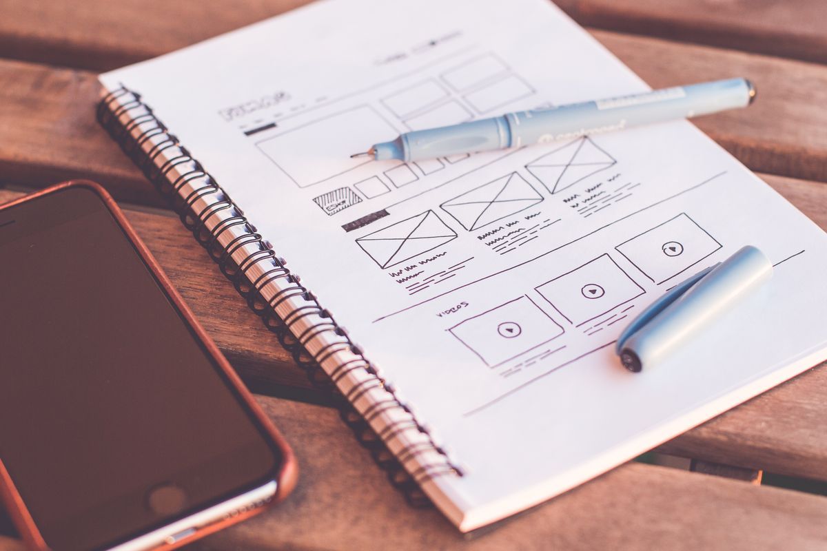 A spiral notebook with hand-drawn website wireframe sketches and a pen, next to a smartphone on a wooden desk