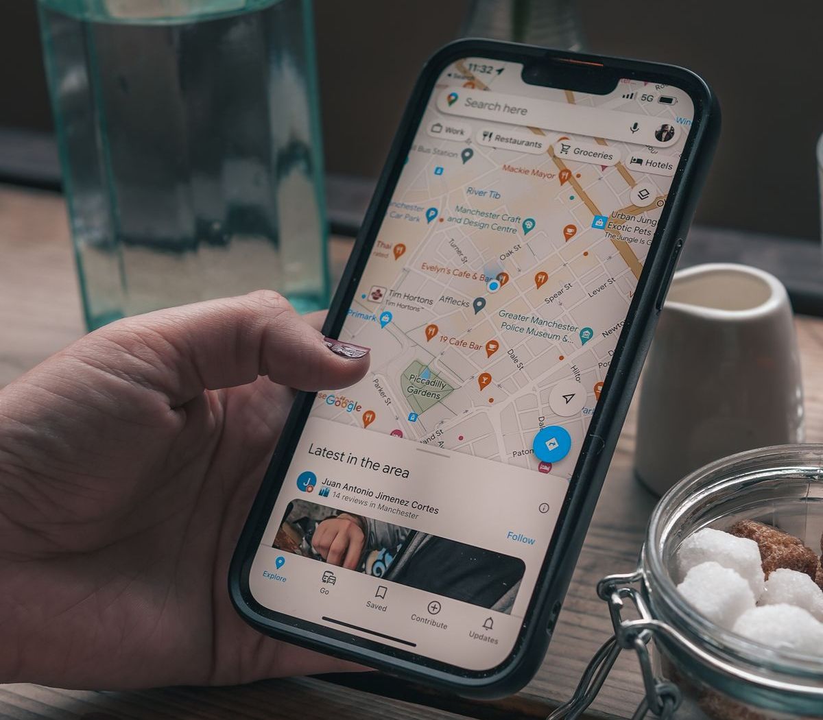 A hand holding a smartphone showing Google Maps with nearby cafés and shops in Manchester, England