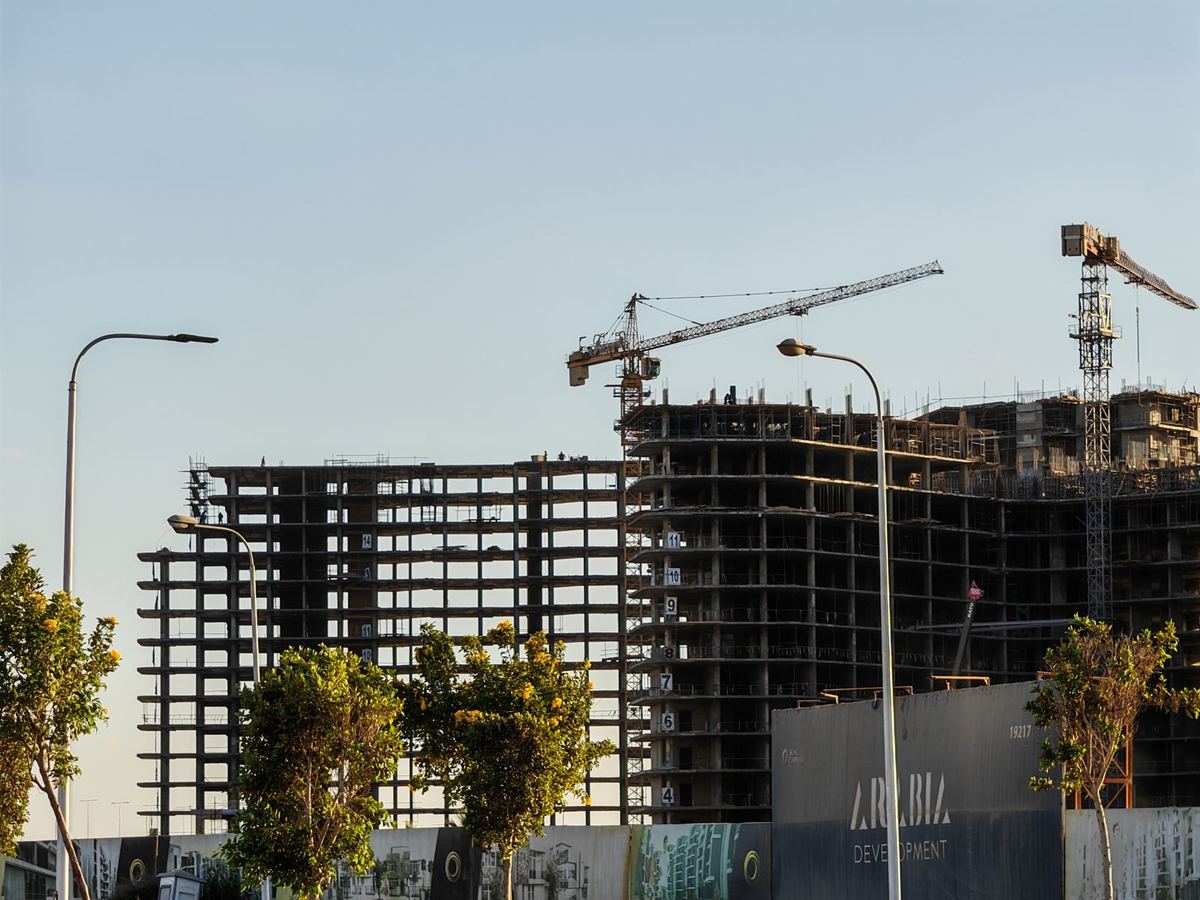 A high-rise building under construction in Dubai, with tower cranes above the concrete frame and trees in the foreground